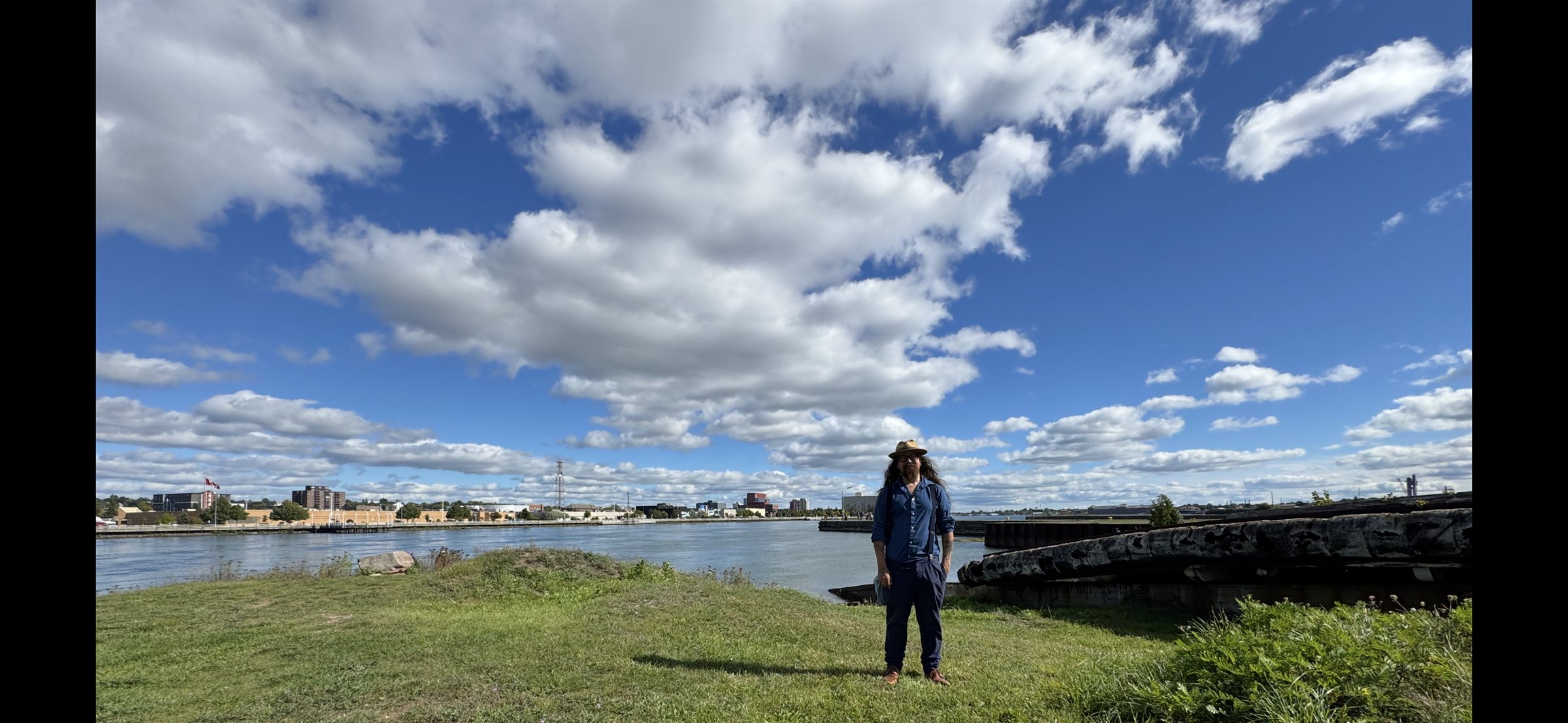 Josh McNally standing beside the waterfront under a wide, cloud-filled blue sky