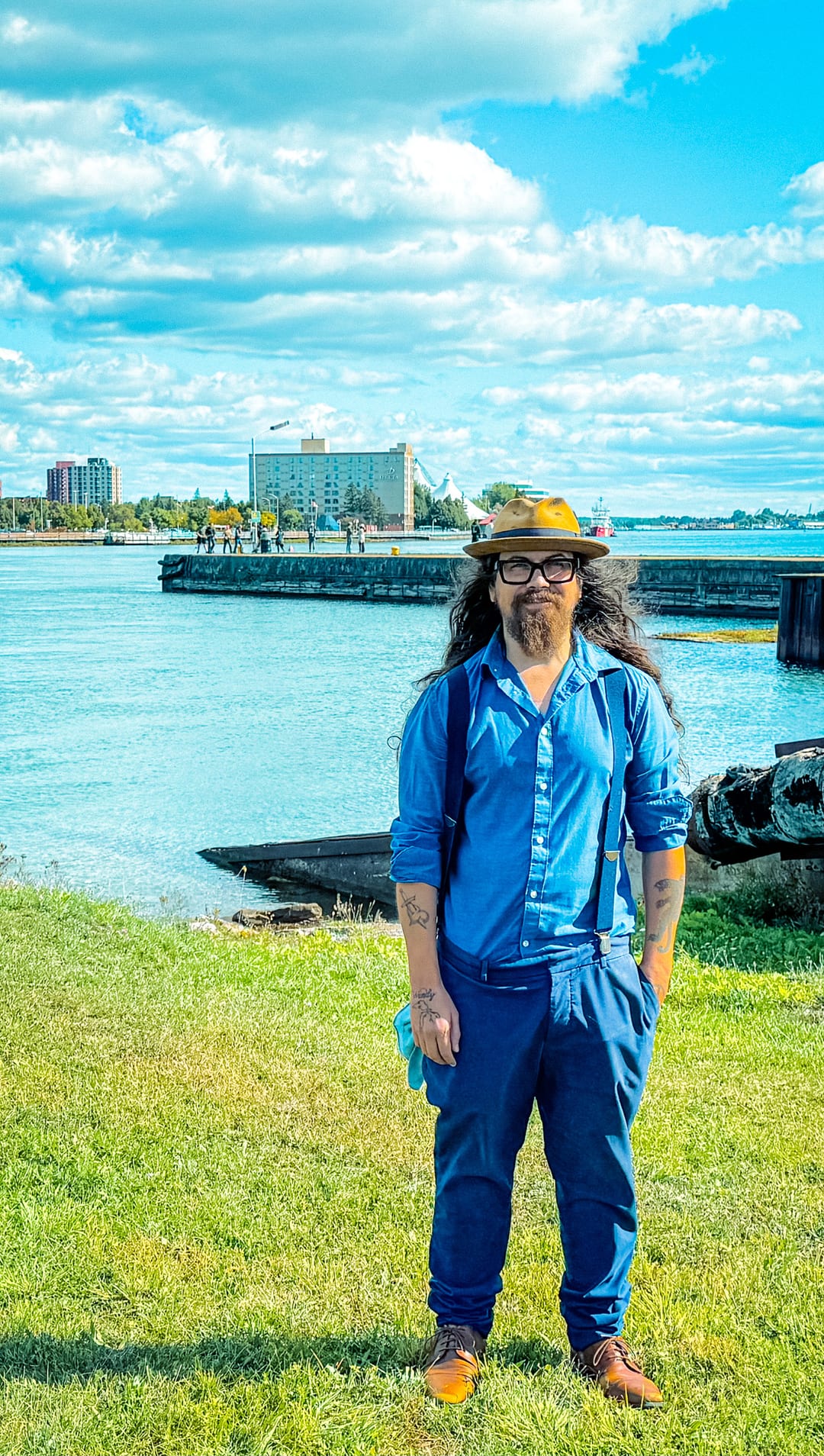 Josh in a blue shirt, brown hat and glasses beside the river, with the waterfront behind him