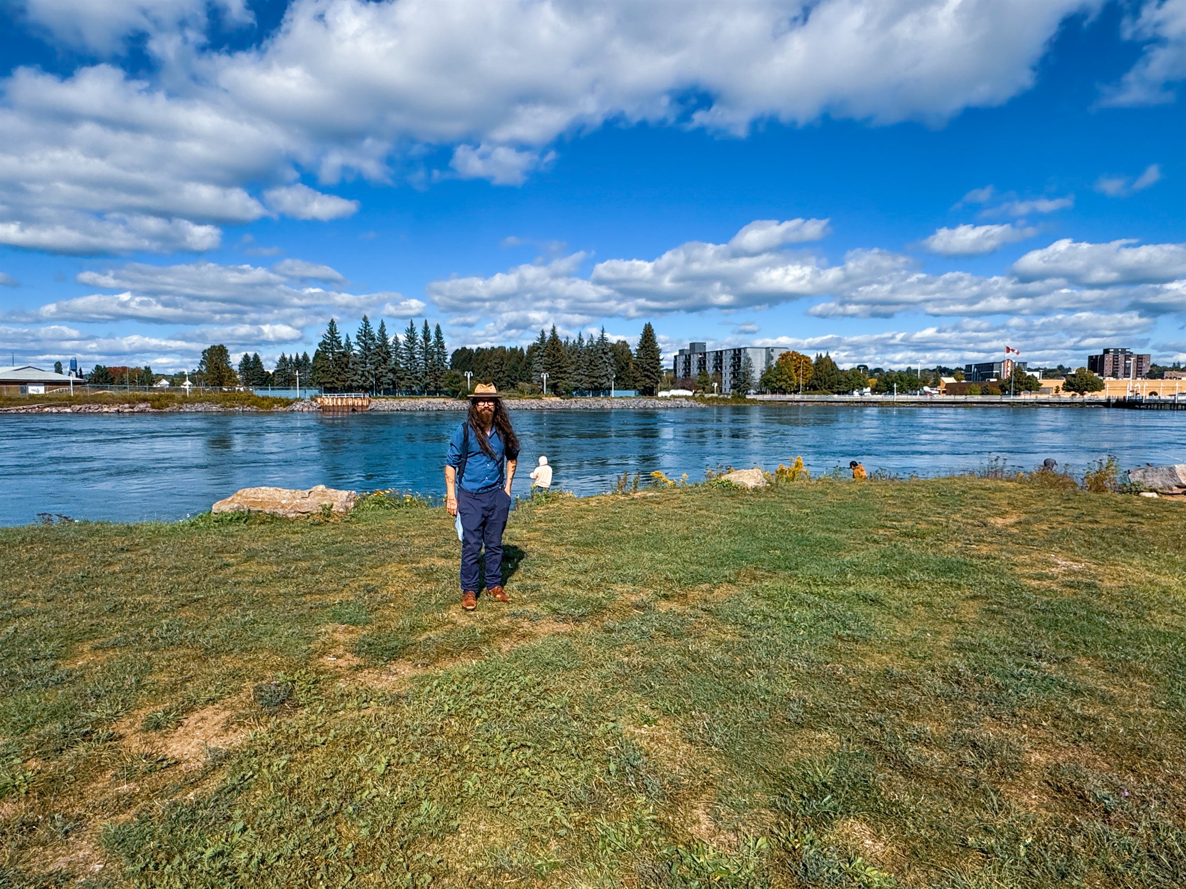 Josh McNally on a grassy riverbank, with the former proposed Gateway site and fish hatchery area across the river behind him